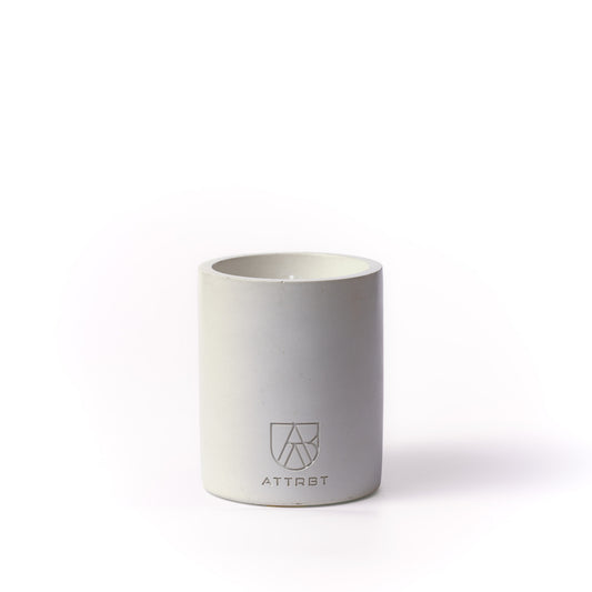 A minimalist one-wick candle in a light grey color cylindrical concrete container, featuring the 'ATTRBT' brand logo on the front, set against a clean white background.
element. concrete candle
